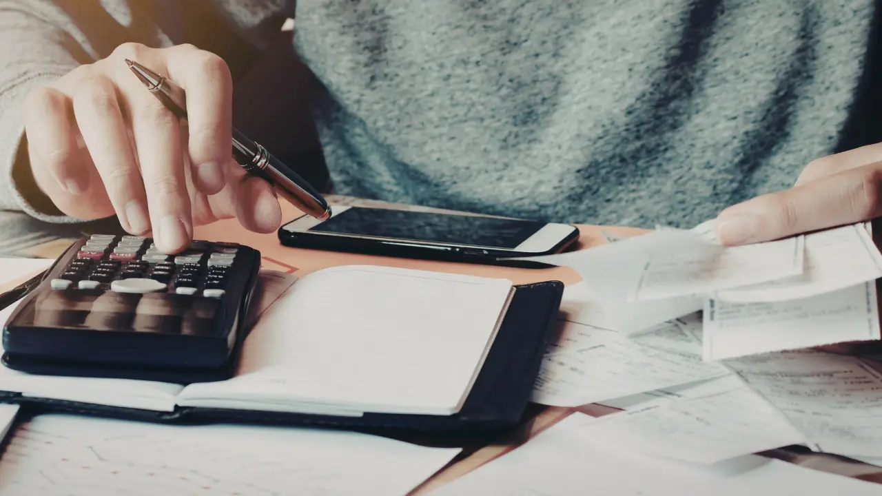 Personne calculant son taux d'endettement avec une calculatrice, entourée de factures et documents financiers, un carnet et un smartphone posés sur le bureau.